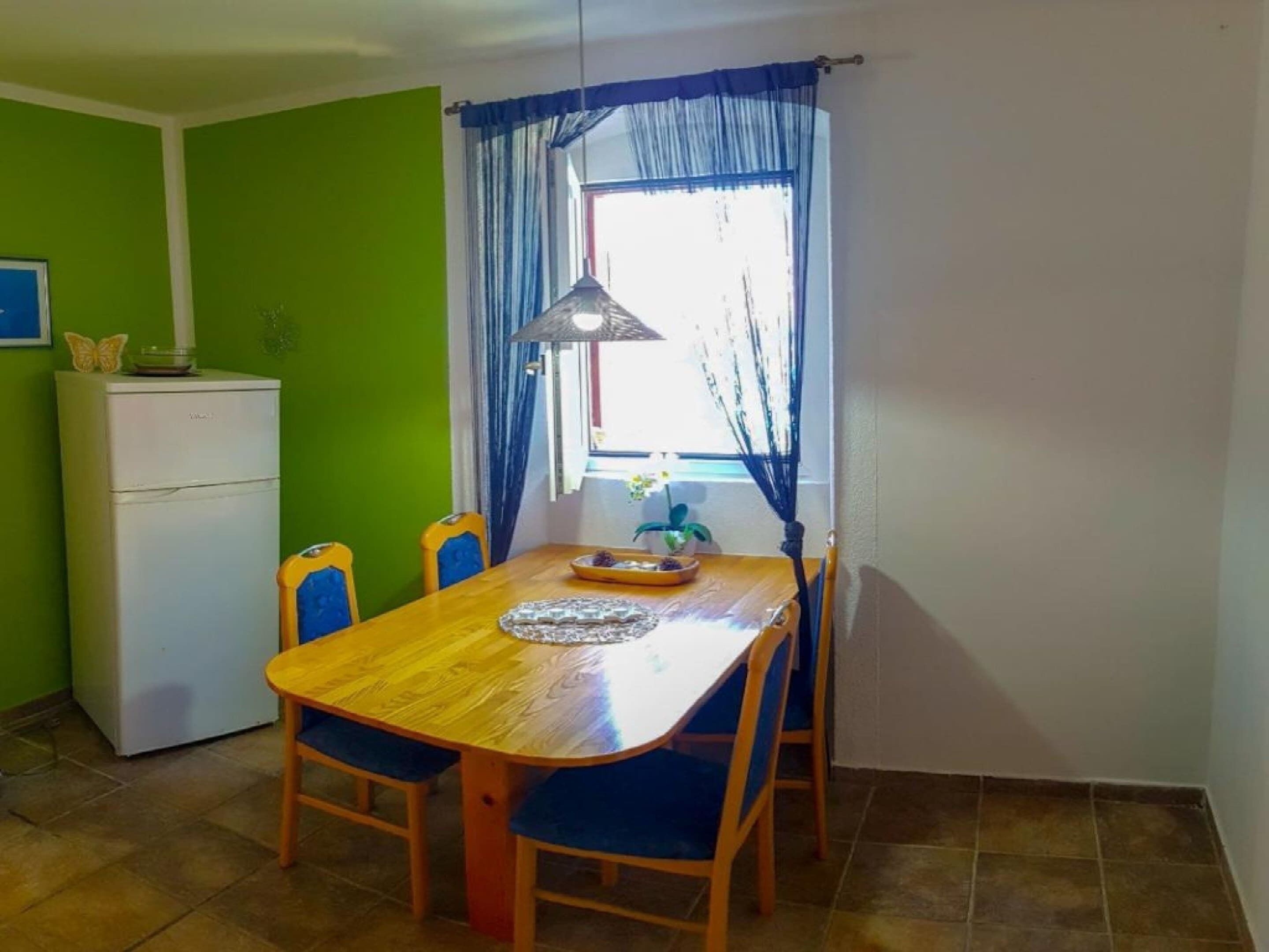 A compact dining area with a wooden table and blue chairs, a small refrigerator, and a window with blue curtains.