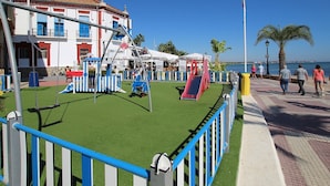 A playground with swings and slides, a fenced area, and a building with red and white walls in the background. - Belvilla by OYO Holiday Home in Los Alcazares (Los Alcazares)