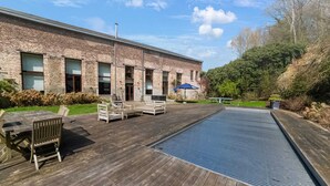 Ett trädäck med utemöbler, en swimmingpool och en byggnad i tegel med fönster.