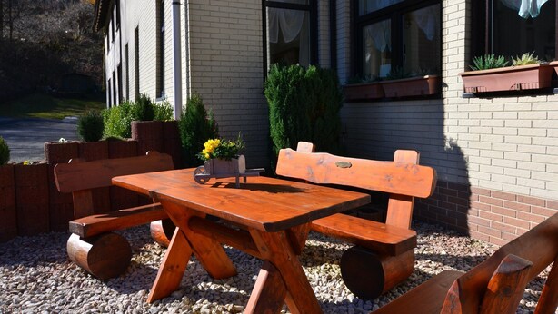 Une table et des bancs en bois, installés à l’extérieur, accompagnés d’un jardinier et de plantes en pot.