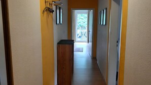 Un couloir étroit avec un banc en bois, une porte donnant sur un espace extérieur et une petite poubelle.