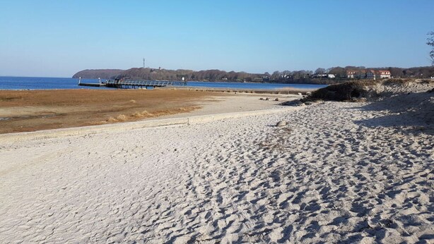 Ein Sandstrand mit einem Steg, der sich ins Wasser erstreckt, ein klarer Himmel und Gebäude im Hintergrund.