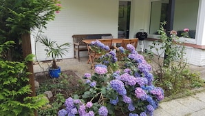Eine Terrasse mit Holztisch und -stühlen, umgeben von einem Garten mit Hortensien und einem Blumentopf.