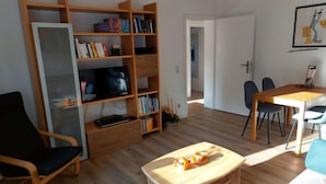 Ein Wohnzimmer mit einem Bücherregal aus Holz, einem Fernseher, einem Esstisch mit Stühlen und einem kleinen Couchtisch.