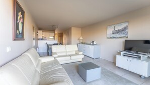 Een moderne woonkamer met een bank, een salontafel, een tv-meubel met een flatscreen televisie, en een keuken in de achtergrond.