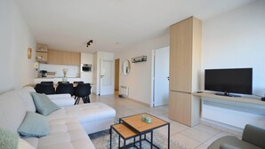 Ein modernes Wohnzimmer mit einer Couch, einem Couchtisch und einem Fernseher. Im Hintergrund ist eine Küchenzeile mit Schränken und einem Kühlschrank zu sehen.