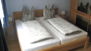 Una cama doble con estructura de madera, dos almohadas con estampado de árbol, una mesita de noche con una lámpara y un cuadro en la pared.