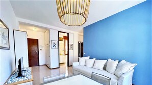 A modern living room with a white sofa, a glass coffee table, and a TV on a stand. There is a blue accent wall and a wooden door leading to another room. - Apartment Near Porto Santa Margherita Beach (Caorle)