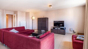 A modern living room with a red sofa, a dark cabinet, a flat-screen TV, and a floor lamp. - Apartment in Koksijde Near the Sea (Koksijde)