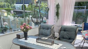 A cozy outdoor seating area with wicker furniture, a table with a vase of flowers, and a view of a garden and buildings. - 8 Person Holiday Home in Hallekis (Hallekis)