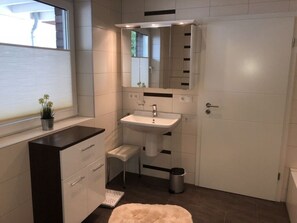 A modern bathroom with a white sink, mirror, and vanity. There is a small trash can and a potted plant on the windowsill. - Holiday Apartment \"dilborner Benden\" (Niederkruechten)