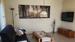 Uma sala de estar com um sofá preto, uma mesa de centro de madeira, um quadro grande com tema de floresta, um abajur, uma televisão e um armário.