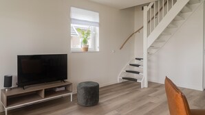 Een moderne woonkamer met een flatscreen televisie op een houten standaard, een grijze poef en een trap met witte leuningen.