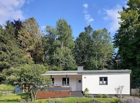 A modern single-story house with a flat roof, a small porch, and a front yard with a wooden deck and a flower bed. - Wamel Modern Retreat (Moehnesee)