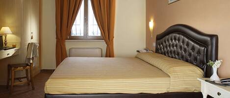 A bedroom with a large bed, a desk with a lamp, a chair, and a window with curtains.