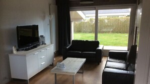 A modern living room with a flat-screen TV, a white cabinet, a black sofa, and a glass coffee table. - Lovely Apartment in Julianadorp (Julianadorp)