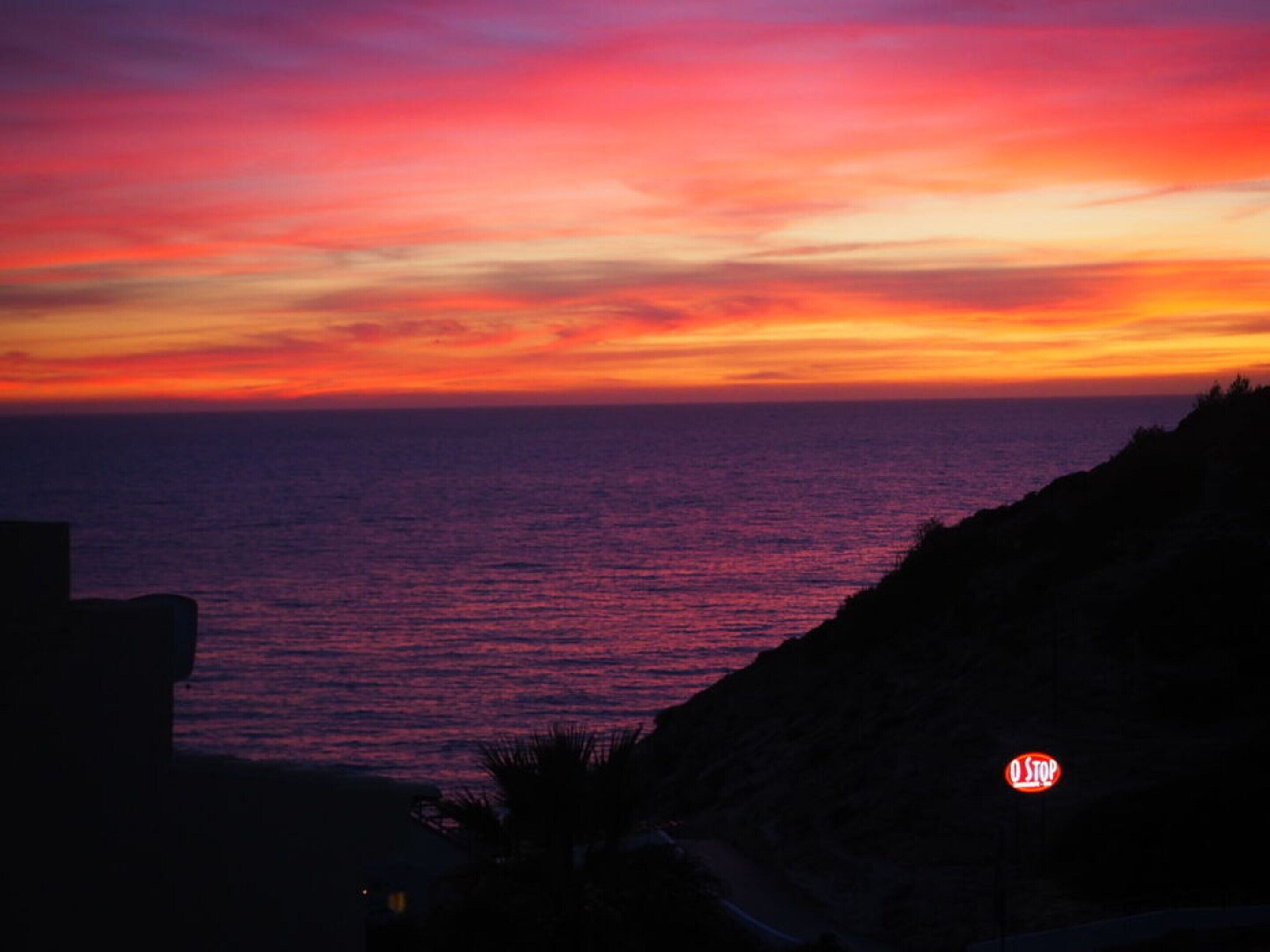 A sunset over the ocean with a rocky coastline and a stop sign visible.