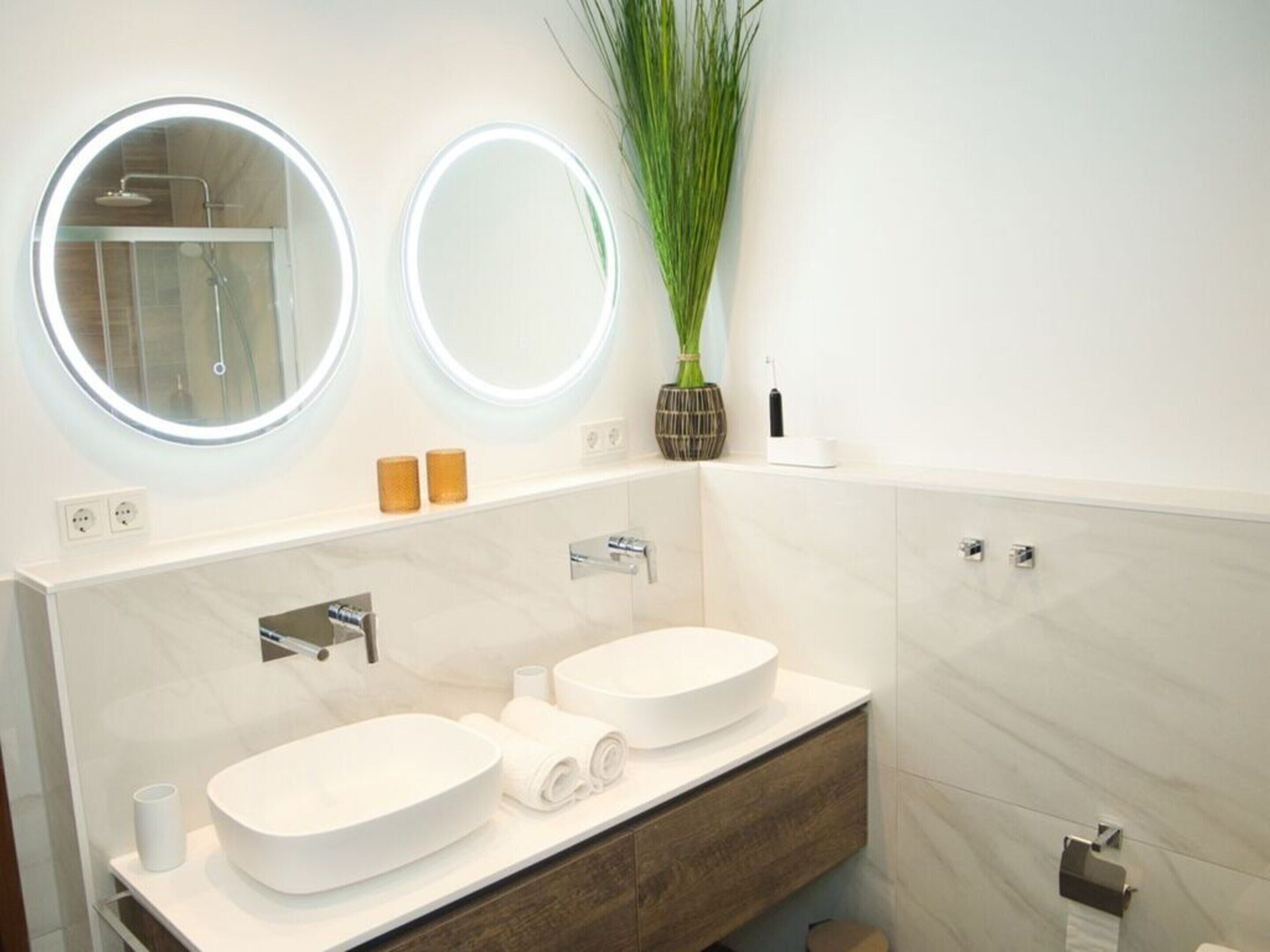 A modern bathroom with two sinks, a shower, and two illuminated mirrors.