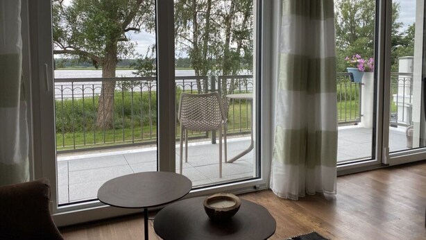 Huoneessa on suuret, liukuvat lasiovet, jotka johtavat parvekkeelle, jolta avautuu näkymä puiden ja vesistön ylle. Huoneessa on pyöreä pöytä, jonka päällä on kulho, ja parvekkeella näkyy tuoli.