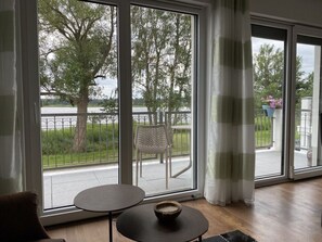 A room with large sliding glass doors leading to a balcony with a view of trees and a body of water. There is a round table with a bowl on it, and a chair is visible on the balcony. - Holiday Apartment Feisneckblick (Waren (Müritz))