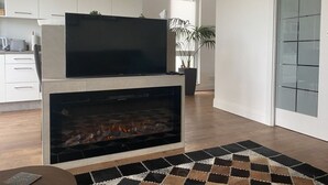 Moderni olohuone, jossa on takka, litteänäyttöinen televisio ja taustalla keittiötila.