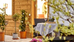 An outdoor dining area with wooden walls, potted plants, and a table set for two with wine glasses and a heart-shaped decoration. - Holiday Apartment in Bavaria (Schoenau am Koenigssee)