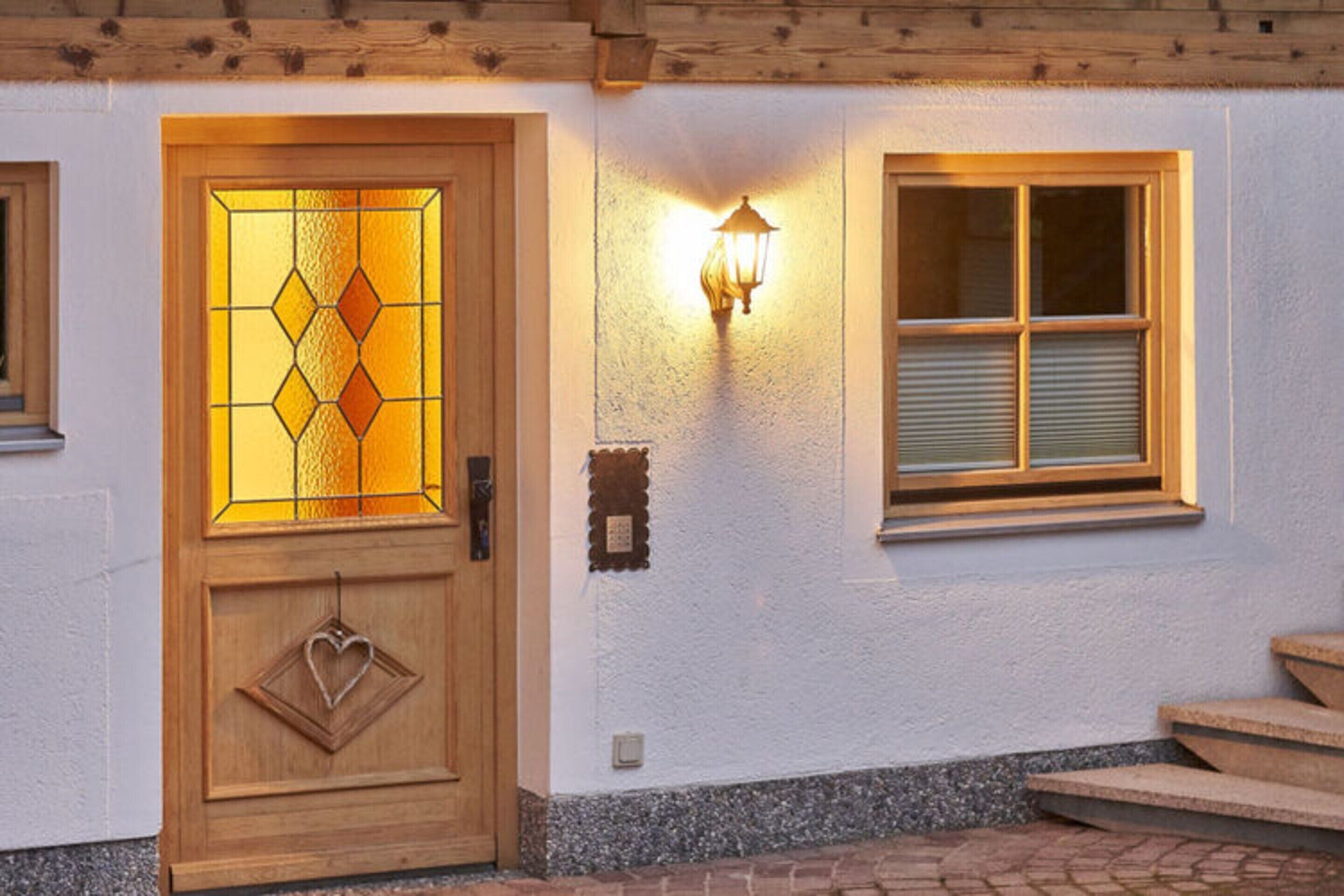 A wooden door with a stained glass panel and a heart-shaped ornament, a wall-mounted light fixture, and a window with blinds.