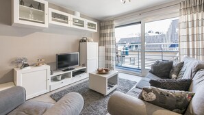 A modern living room with a flat-screen TV, white cabinetry, and a large window with a view of buildings. - Apartment Near the Grand Place (Blankenberge)