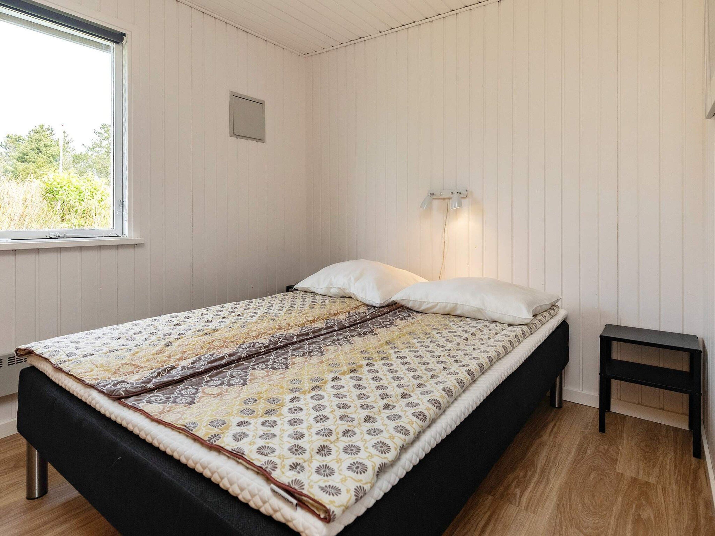 Un lit simple avec une couvre-lit à motifs, une table de chevet, un luminaire fixé au mur et une fenêtre donnant sur un paysage verdoyant.