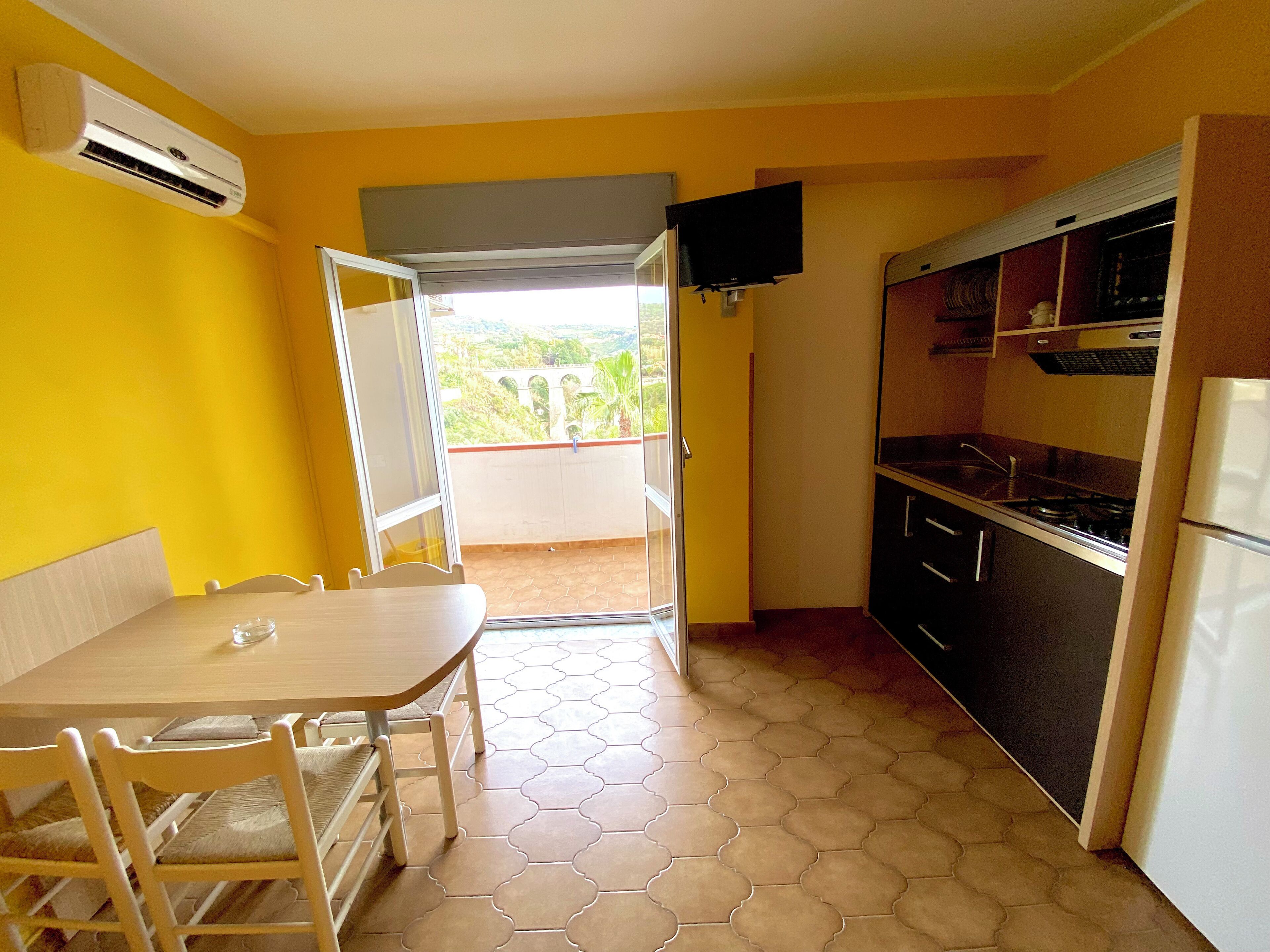 A compact kitchen with a dining area, a TV mounted on the wall, and a view through an open door.