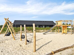 A playground with swings, a slide, and a wooden climbing structure on a sandy area with a building in the background.