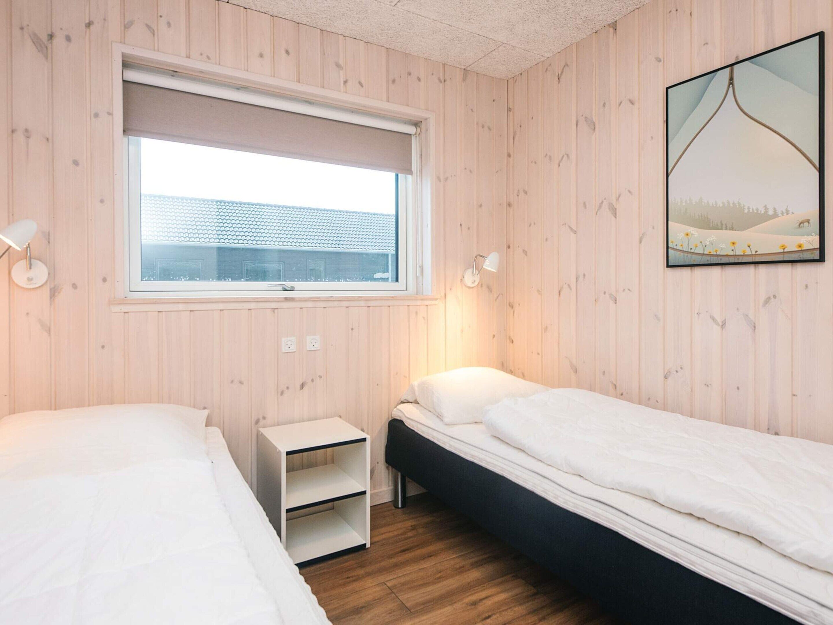 A bedroom with two beds, a window, a bedside table, and a wall-mounted lamp.