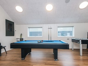 A pool table with a blue felt surface, a foosball table, and two windows with blinds.