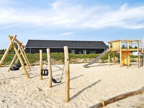Ein Spielplatz mit Schaukeln und einer Rutsche, ein Gebäude mit dunklem Dach und ein sandiger Bereich.