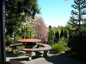 A stone outdoor seating area with a central table and four benches, surrounded by lush greenery and flowering trees.