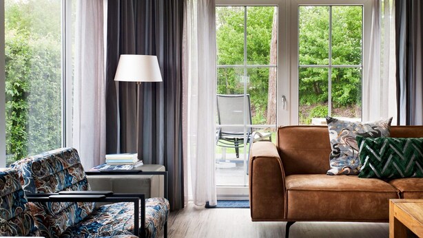A living room with a sofa, a coffee table, and a lamp. There is a view of greenery outside through large windows. - Nice Home in Earnewald With Whirlpool and Sauna (Earnewald)