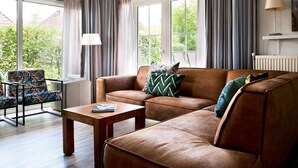 A modern living room with a brown leather sofa, a wooden coffee table, and a patterned armchair. Large windows with curtains allow natural light and views of greenery outside. - Nice Home in Earnewald With Whirlpool and Sauna (Earnewald)