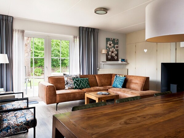A modern living room with a brown leather sofa, a wooden coffee table, and a dining table. There is a window with curtains, a painting on the wall, and a television on the right.