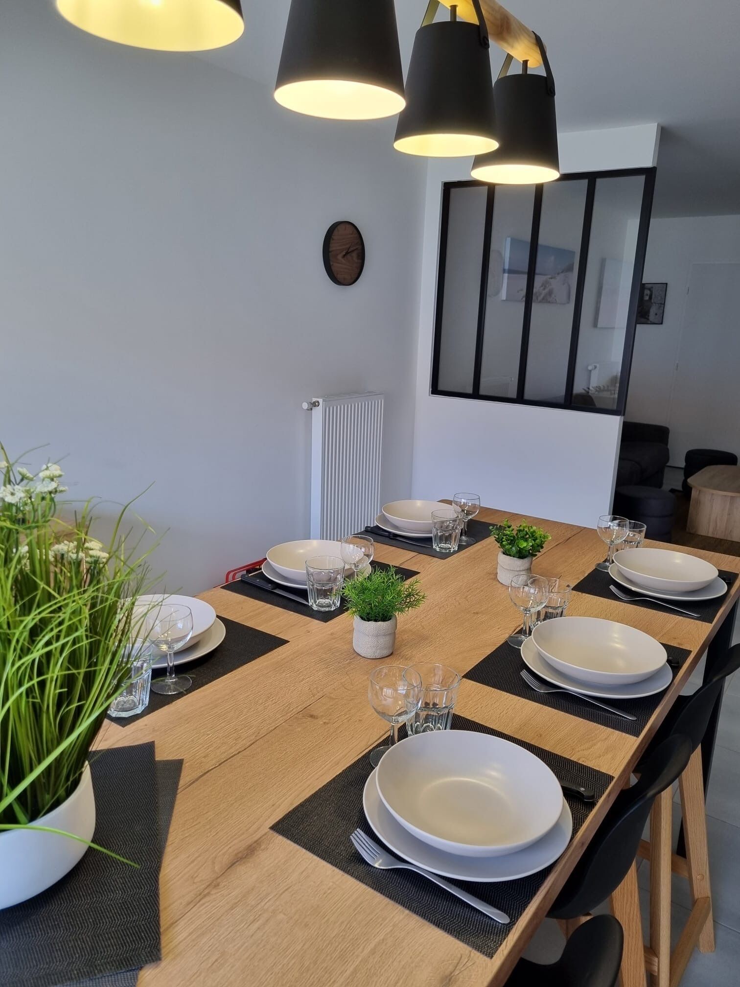 A dining area with a long wooden table set for four, black placemats, white plates, and clear glasses. There is a small potted plant on the table, and a clock on the wall.