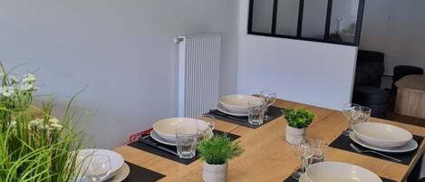 A dining area with a long wooden table set for four, black placemats, white plates, and clear glasses. There is a small potted plant on the table, and a clock on the wall.