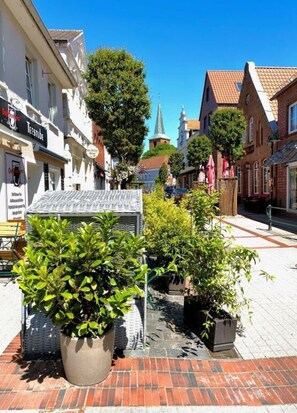 Eine enge Straße, gesäumt von traditionellen europäischen Gebäuden, mit einem Kirchturm im Hintergrund und Pflanzkübeln am Gehweg.