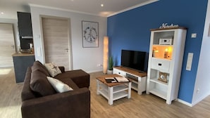 A modern living room with a brown sofa, a TV on a wooden stand, and a white entertainment unit against a blue wall. - Near the North Sea Modern Retreat (Esens)