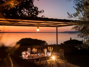 Een terras met een eettafel gedekt voor twee personen, onder een pergola met een hangende lamp, met uitzicht op een rustig wateroppervlak en verre heuvels.
