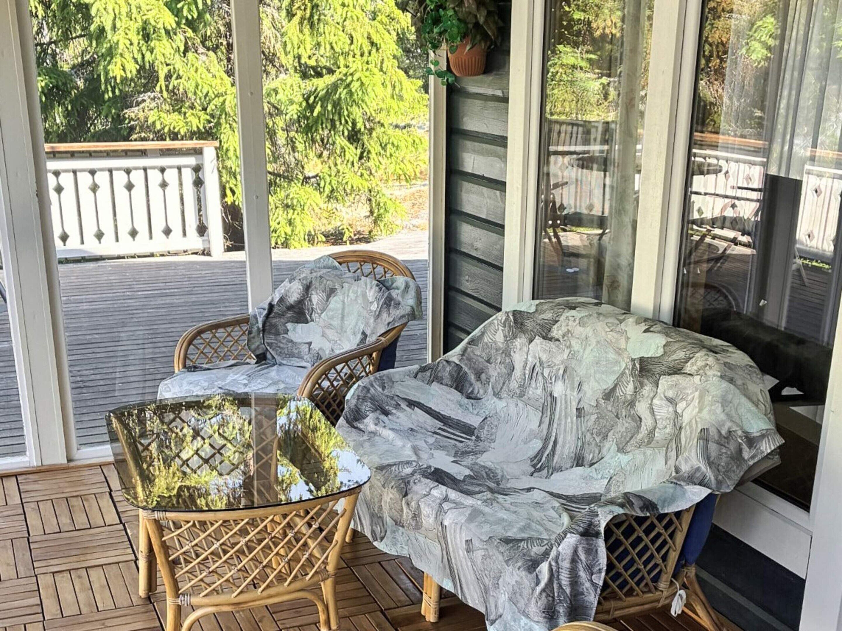 A covered outdoor seating area with wicker chairs and a glass table, overlooking a wooden deck and greenery.