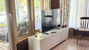 A room with a TV on a white cabinet, a chair, and a window with floral curtains. - 7 Person Holiday Home in Grisslehamn (Grisslehamn)