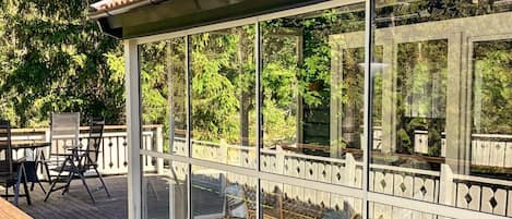 A covered outdoor deck with glass walls, wooden furniture, and a railing.