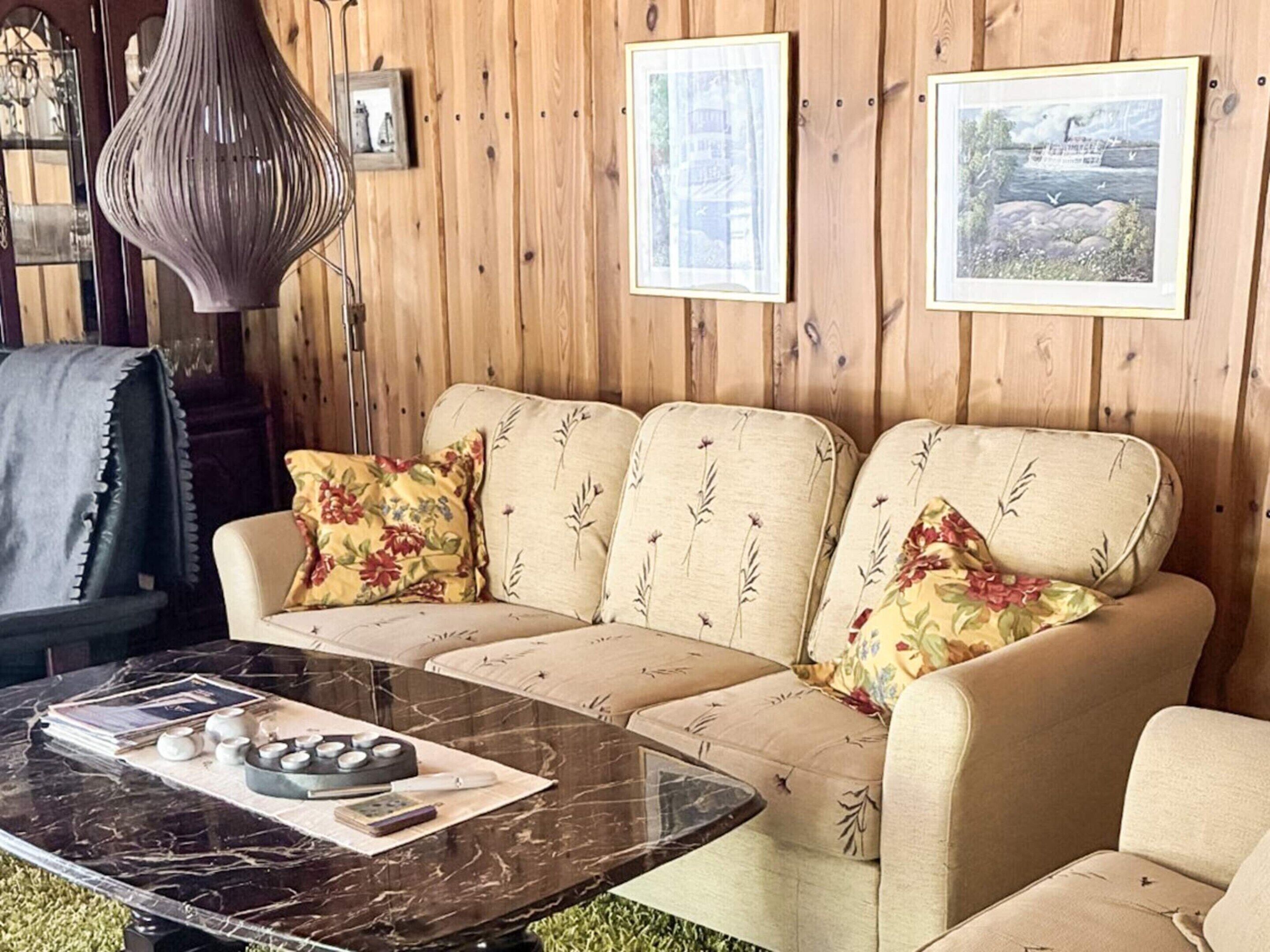 A cozy room with a wooden wall, a sofa with floral cushions, a marble coffee table, and framed pictures.