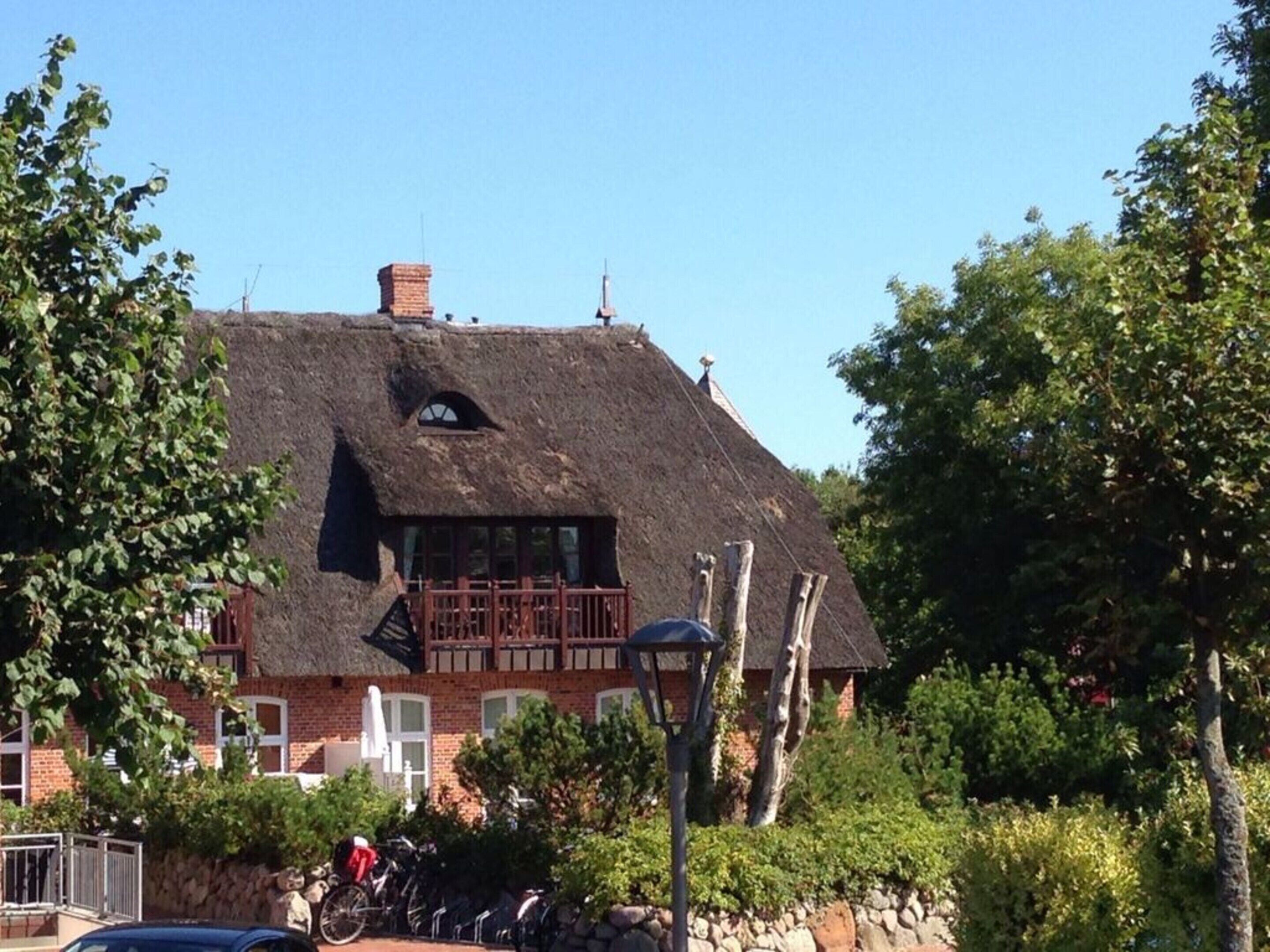 Ein traditionelles Gebäude mit einem Reetdach und einem Balkon, umgeben von viel Grün.