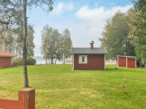 A small red wooden building with a window, surrounded by a grassy field and trees, with a body of water in the background. - 3 Person Holiday Home in Grimstorp (Grimstorp)