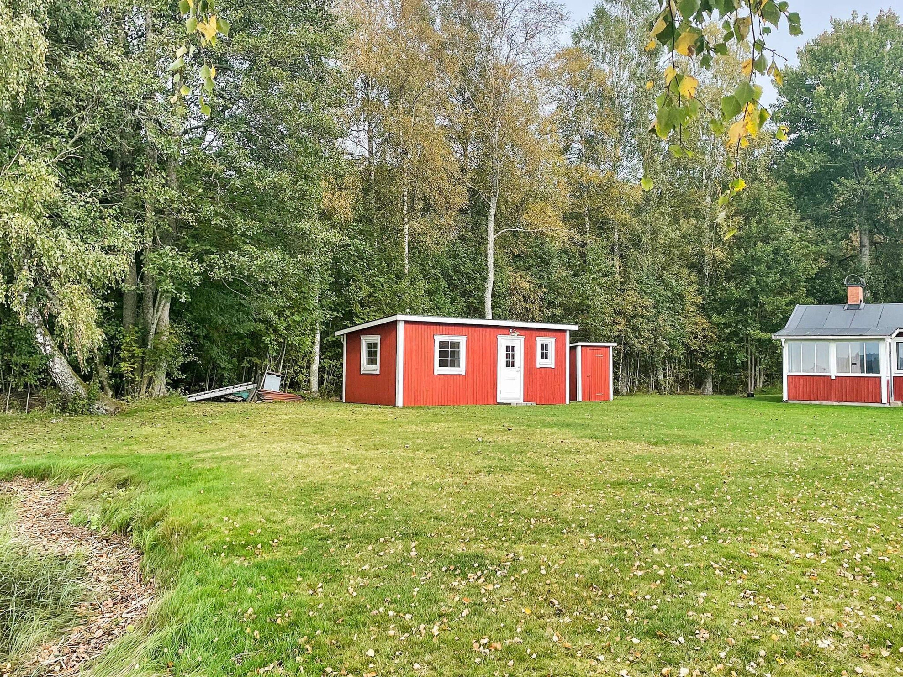 A red wooden cabin with a white door and windows, surrounded by a grassy field and dense trees.